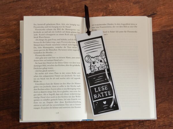 Lesezeichen auf Holzhintergrund: Weißes Papier, schwarzer Linoldruck. Das Motiv ist eine Ratte, die hinter einem Buchrücken hervorschaut, auf dem "Leseratte" steht. Das Lesezeichen wird durch ein schwarzes Satinband ergänzt.