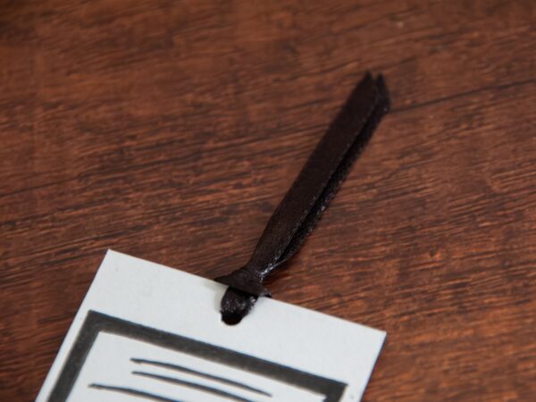Lesezeichen auf Holzhintergrund: Weißes Papier, schwarzer Linoldruck. Das Motiv ist eine Ratte, die hinter einem Buchrücken hervorschaut, auf dem "Leseratte" steht. Das Lesezeichen wird durch ein schwarzes Satinband ergänzt.