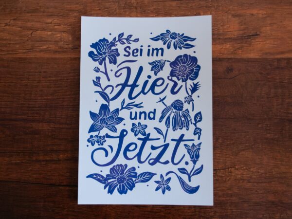 Linoldruck in Din A4: Ein lila Schriftzug "Sei im Hier und Jetzt" umgeben von Blumen und Blättern in einem kräftigen, dunklen Lila