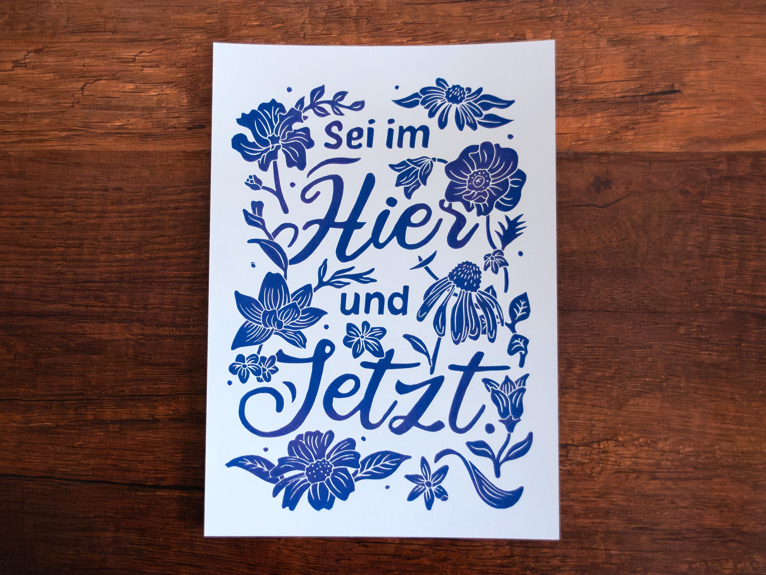 Linoldruck in Din A4: Ein lila Schriftzug "Sei im Hier und Jetzt" umgeben von Blumen und Blättern in einem kräftigen, dunklen Lila
