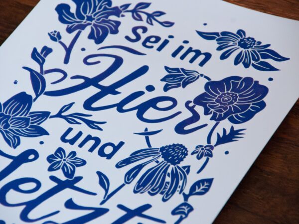 Linoldruck in Din A4: Ein lila Schriftzug "Sei im Hier und Jetzt" umgeben von Blumen und Blättern in einem kräftigen, dunklen Lila