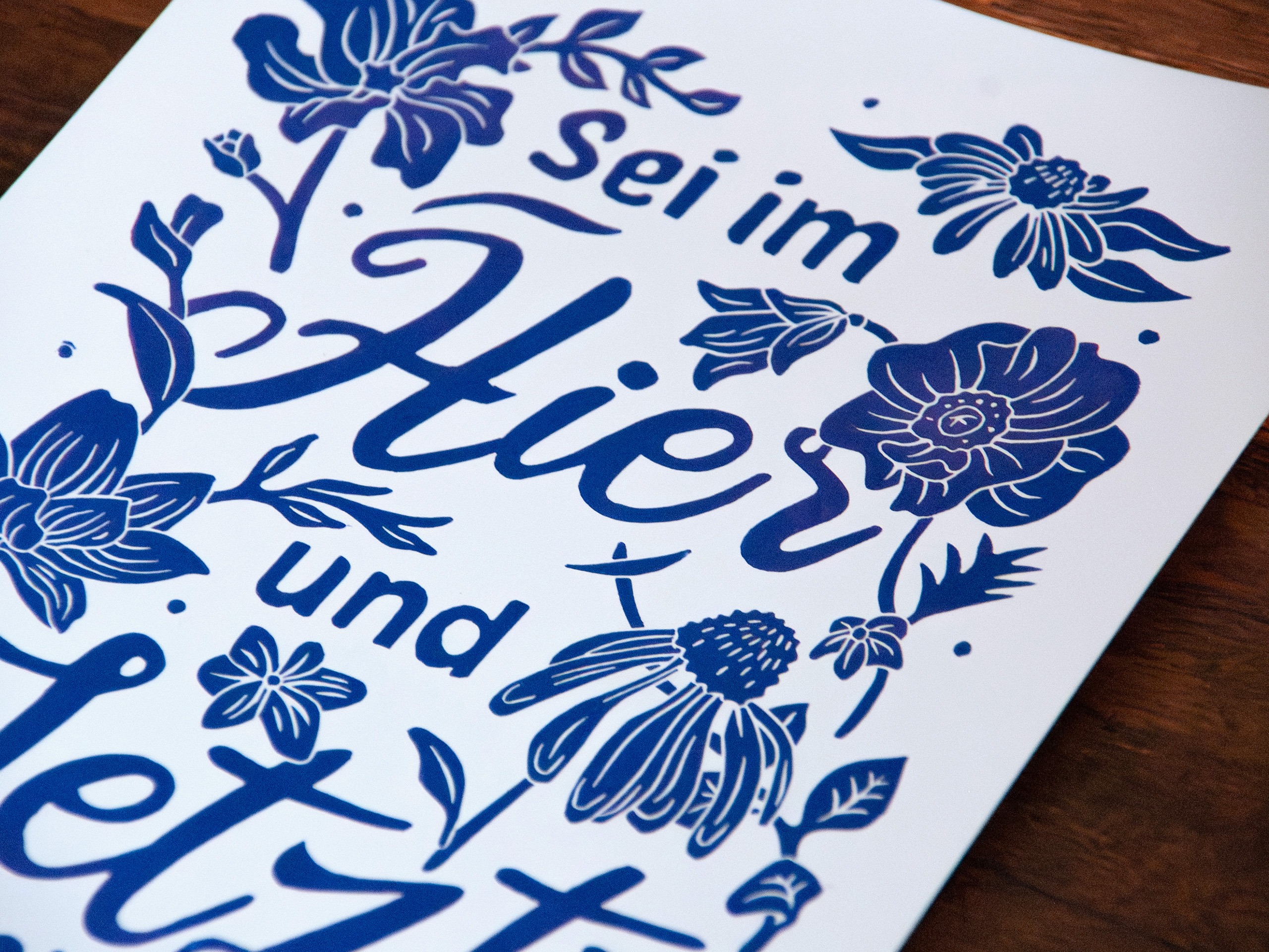 Linoldruck in Din A4: Ein lila Schriftzug "Sei im Hier und Jetzt" umgeben von Blumen und Blättern in einem kräftigen, dunklen Lila