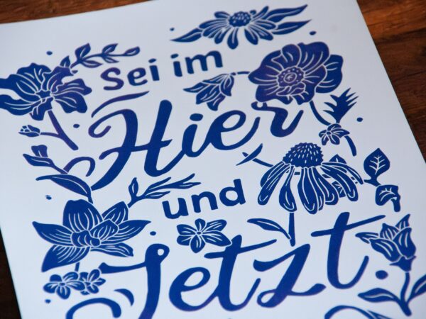 Linoldruck in Din A4: Ein lila Schriftzug "Sei im Hier und Jetzt" umgeben von Blumen und Blättern in einem kräftigen, dunklen Lila