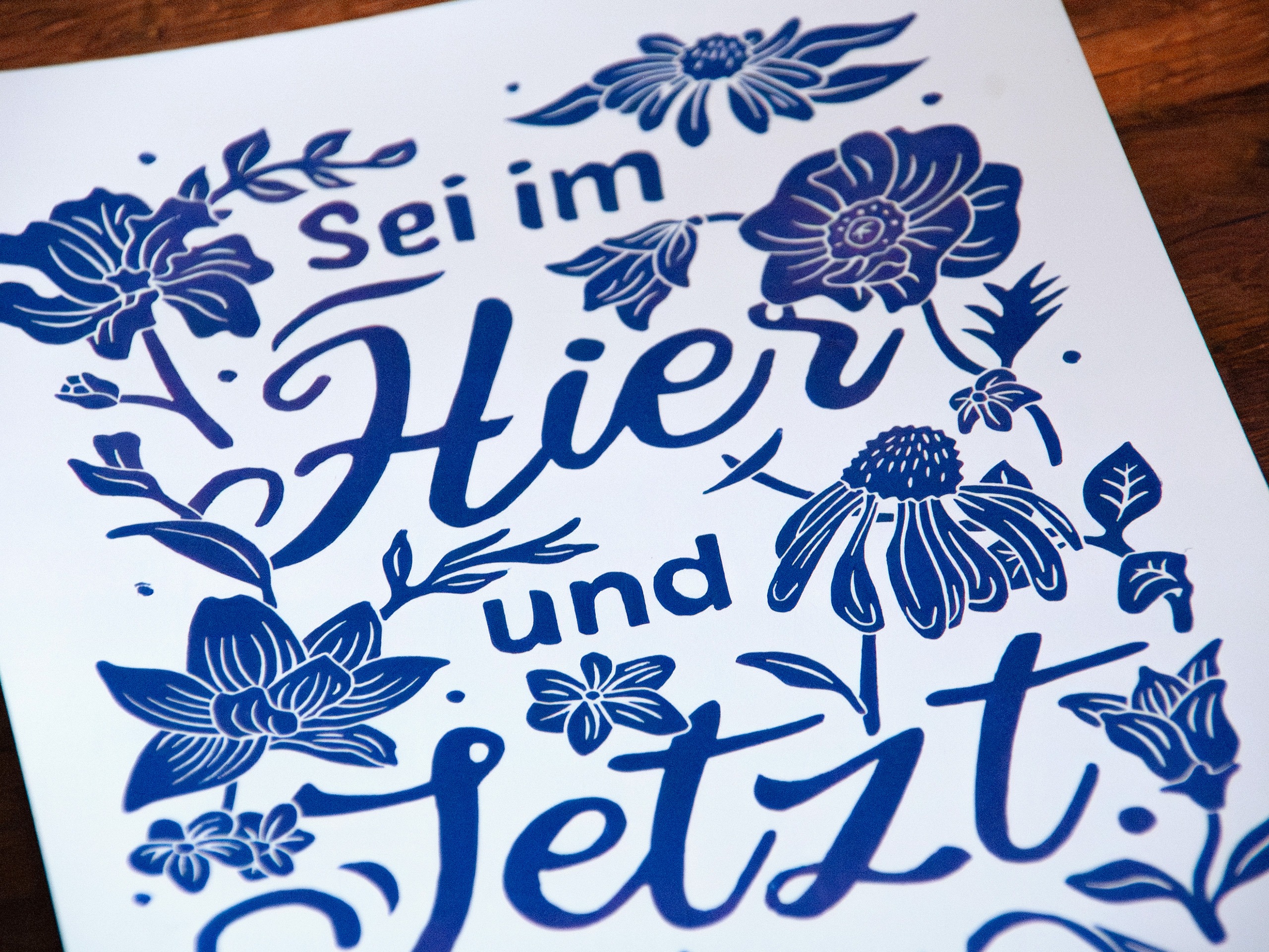 Linoldruck in Din A4: Ein lila Schriftzug "Sei im Hier und Jetzt" umgeben von Blumen und Blättern in einem kräftigen, dunklen Lila