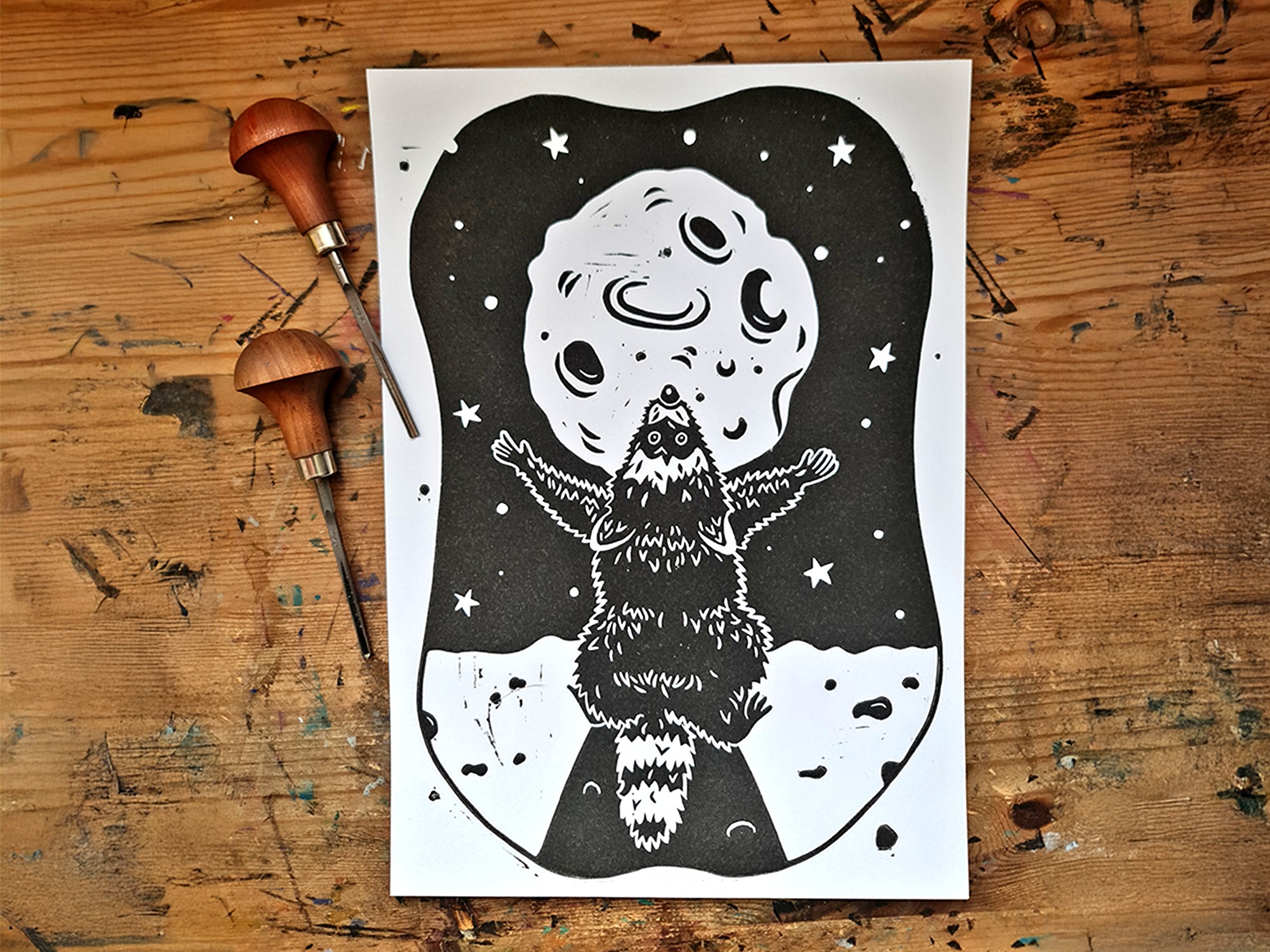 Ein Linoldruck mit einem schwarzen Waschbären, der den Mond anbetet auf einer Holzfläche