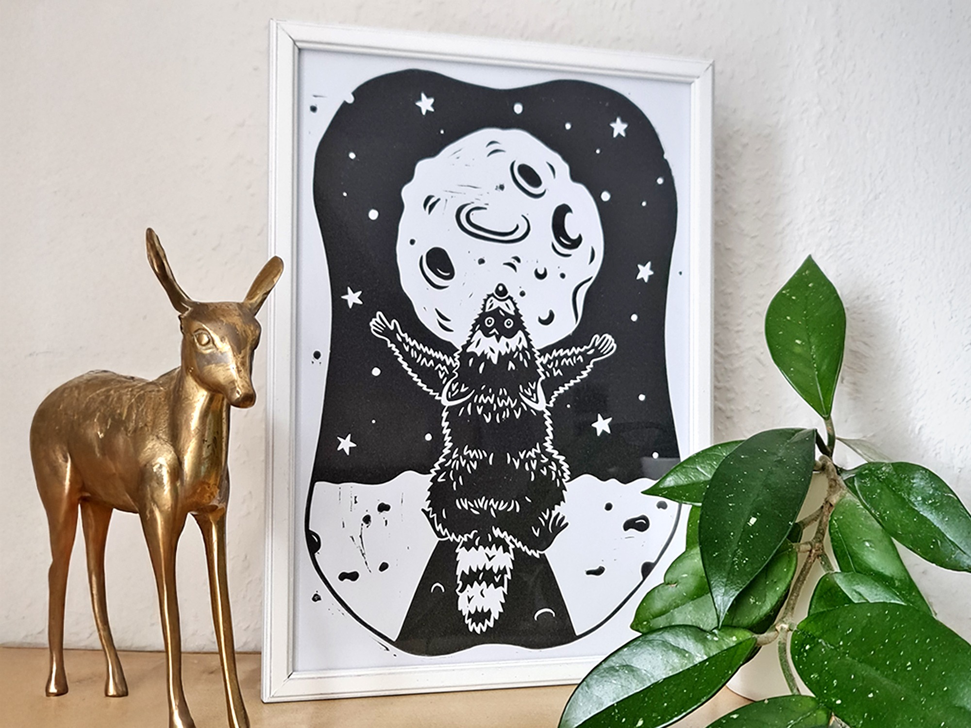Ein Linoldruck mit einem schwarzen Waschbären, der den Mond anbetet auf einer Holzfläche