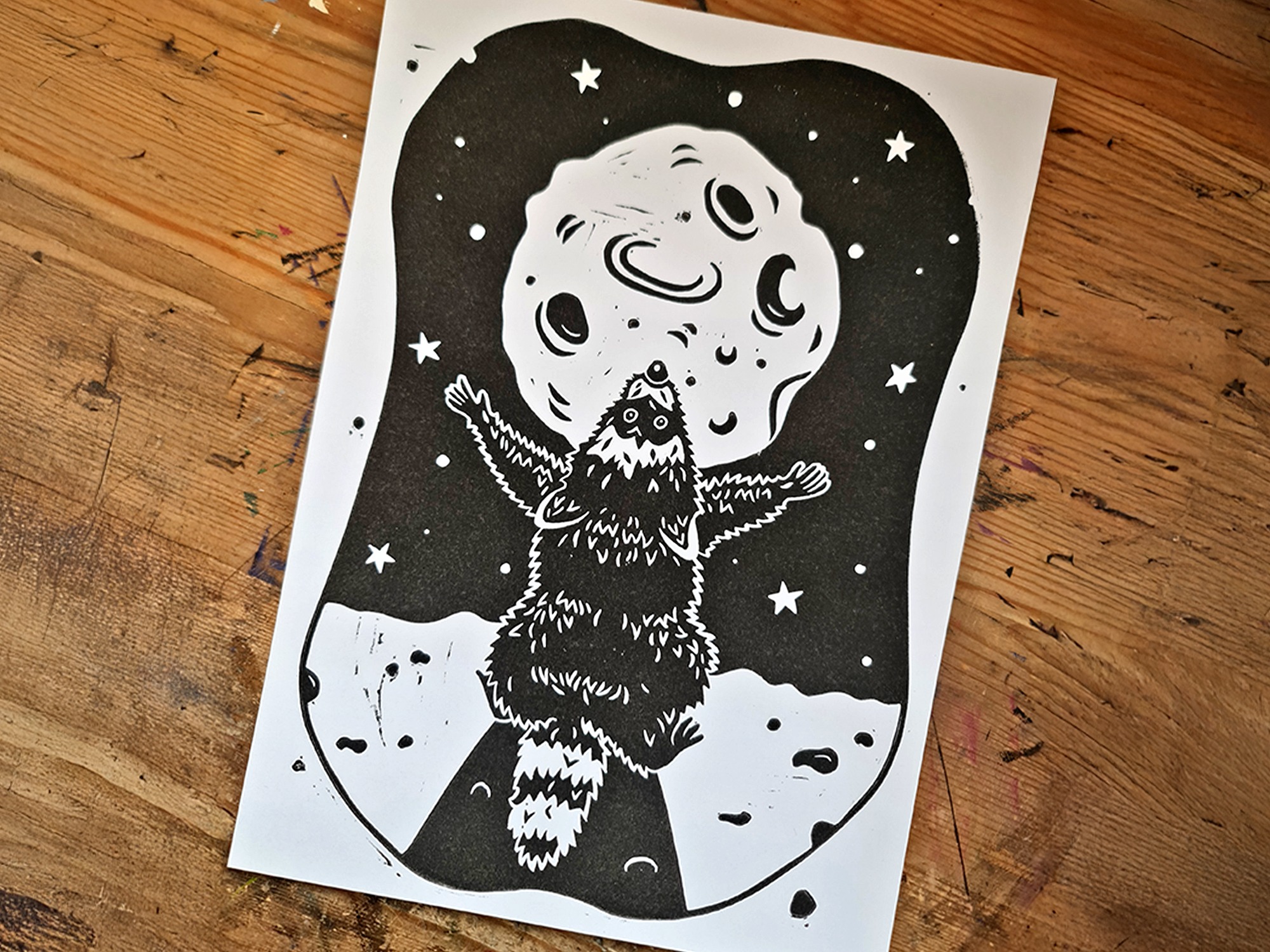 Ein Linoldruck mit einem schwarzen Waschbären, der den Mond anbetet auf einer Holzfläche